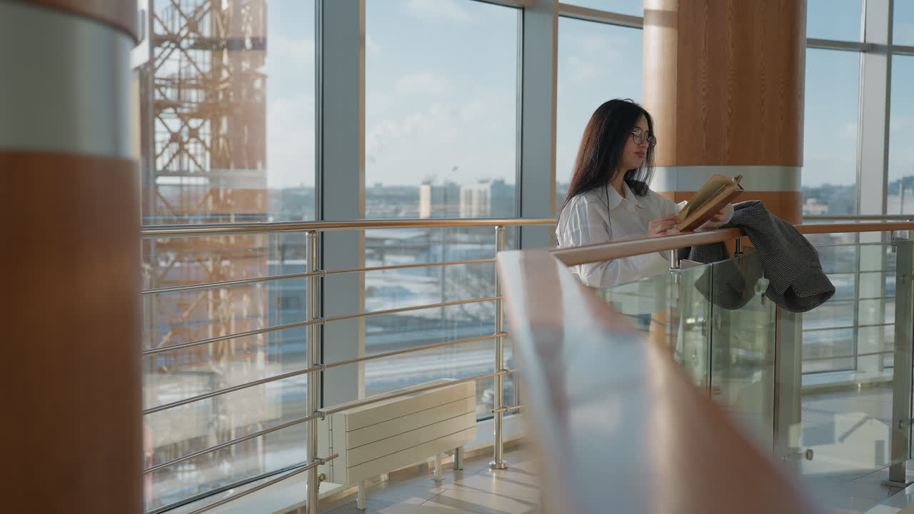 Student lady with long dark hair and glasses reads book with warm smile, leaning by railing inside bright modern building with large glass windows and view of urban cityscape with moving traffic