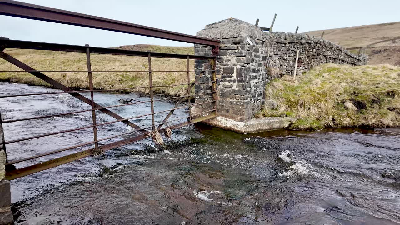 Cancello di metallo arrugginito che attraversa un ruscello che scorre dal bacino idrico di Burnhope, accanto a un vecchio muro di pietra.