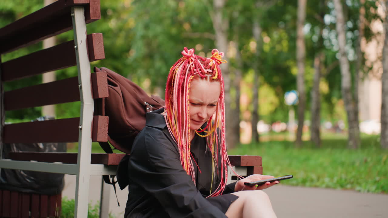 white woman with pink dreadlocks scrolling phone screen while seated on park bench, slight smile and relaxed shoulders, backpack beside her, sunlit birch trees, subtle shift from distress