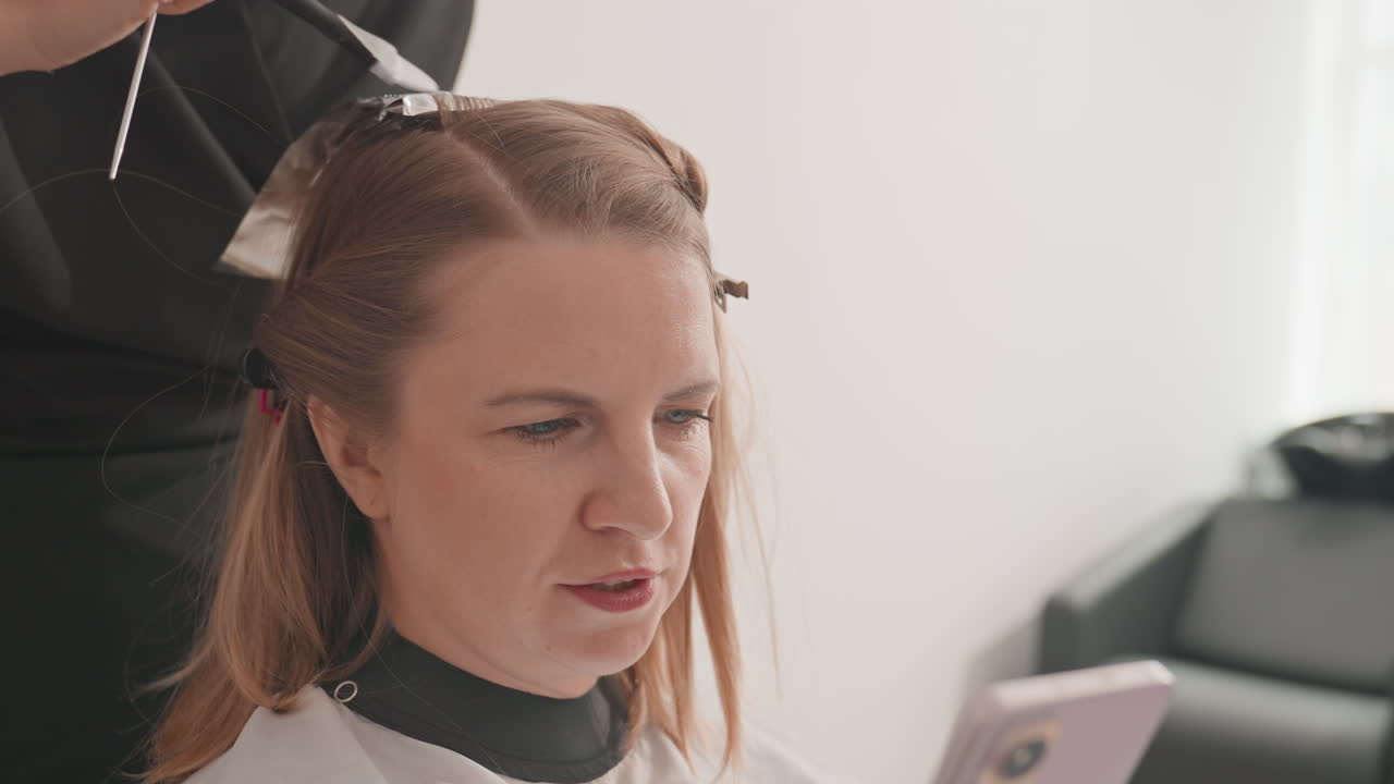 Kaukasisk kvinne i salong leser på telefonen, stylist påfører farge med folie. Klienten sitter med cape og scroller på smarttelefonen, fokusert uttrykk, subtilt naturlig lys, moderne minimalistisk interiør
