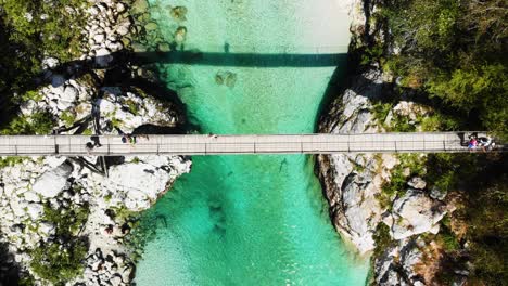 top-down drone view of a suspension bridge over slovenia's soca river with crystal-clear turquoise water, visible stones, and people crossing