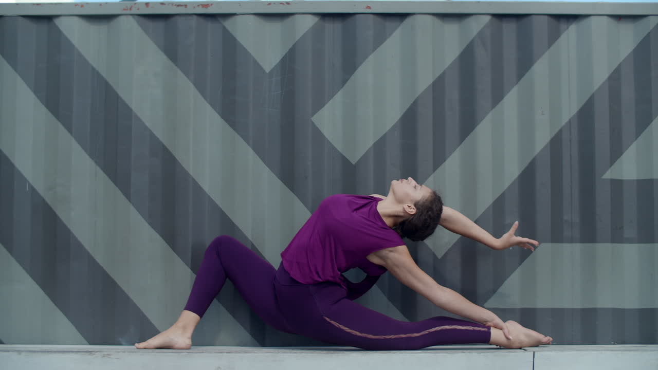 Woman doing yoga split pose outdoors near a gray corrugated metal wall