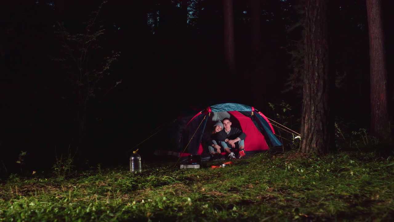 Siblings seated together inside tent during nighttime camping in quiet forest with scattered camping items nearby, surrounded by trees and glowing light from tent