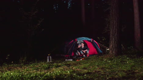 siblings seated together inside tent during nighttime camping in quiet forest with scattered camping items nearby, surrounded by trees and glowing light from tent