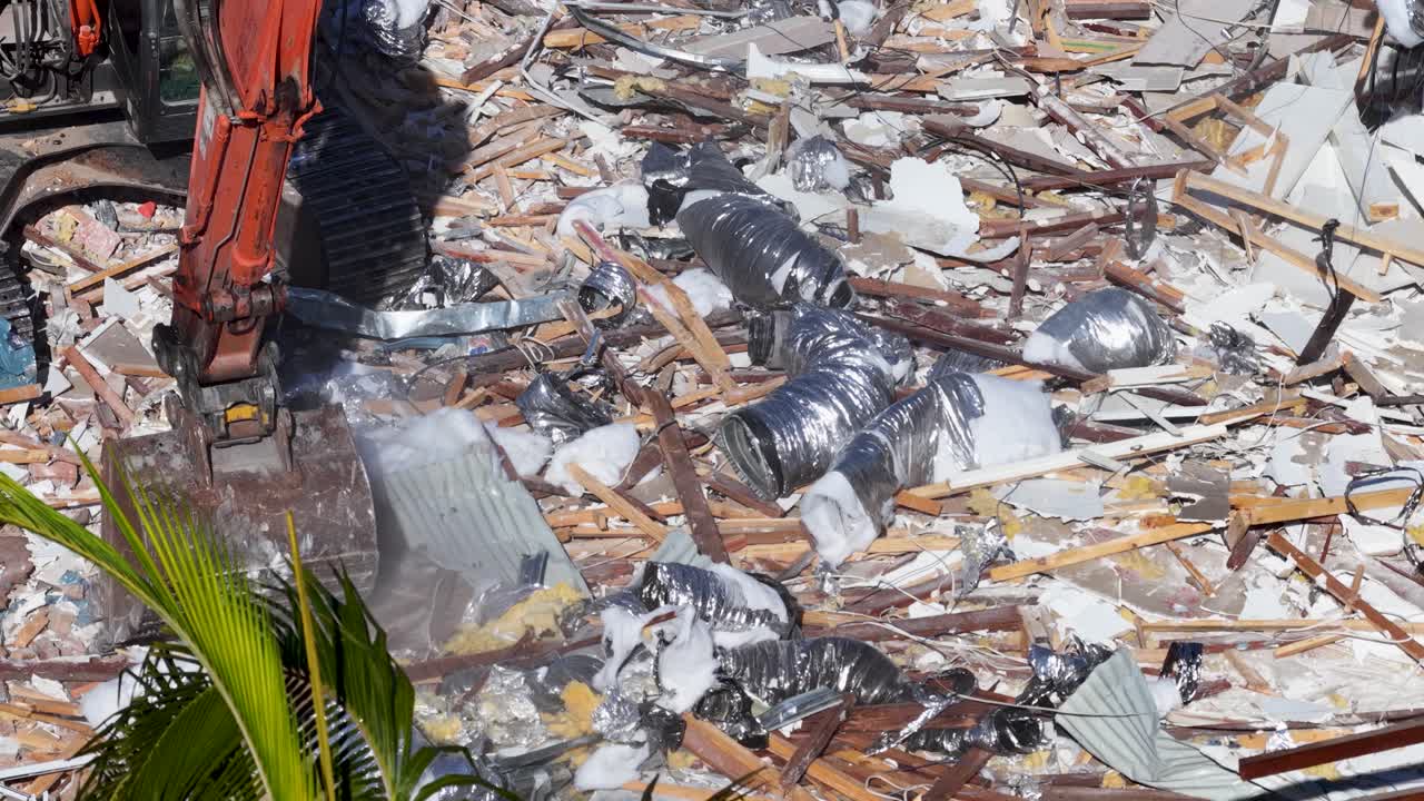 An orange excavator moves rubble and debris from a demolished building in bright daylight, captured in a steady aerial shot over a construction zone