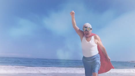 Animation-of-clouds-over-happy-senior-caucasian-man-wearing-superhero-costume-on-beach