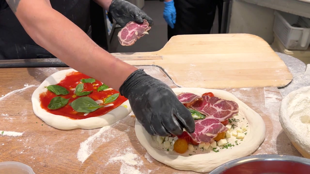Chef con guanti neri posiziona il prosciutto sull'impasto della pizza con salsa di pomodoro e basilico fresco.