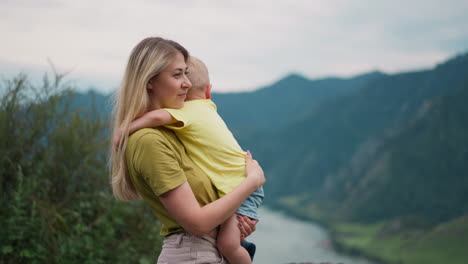 Cheerful-woman-cuddles-boy-against-river-and-mountains