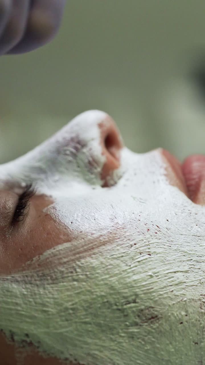 Beautician puts a white mask on the female client's face. Skin care and rejuvenation in modern beauty salon. Side view close up. Vertical video
