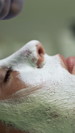 beautician puts a white mask on the female client's face. skin care and rejuvenation in modern beauty salon. side view close up. vertical video