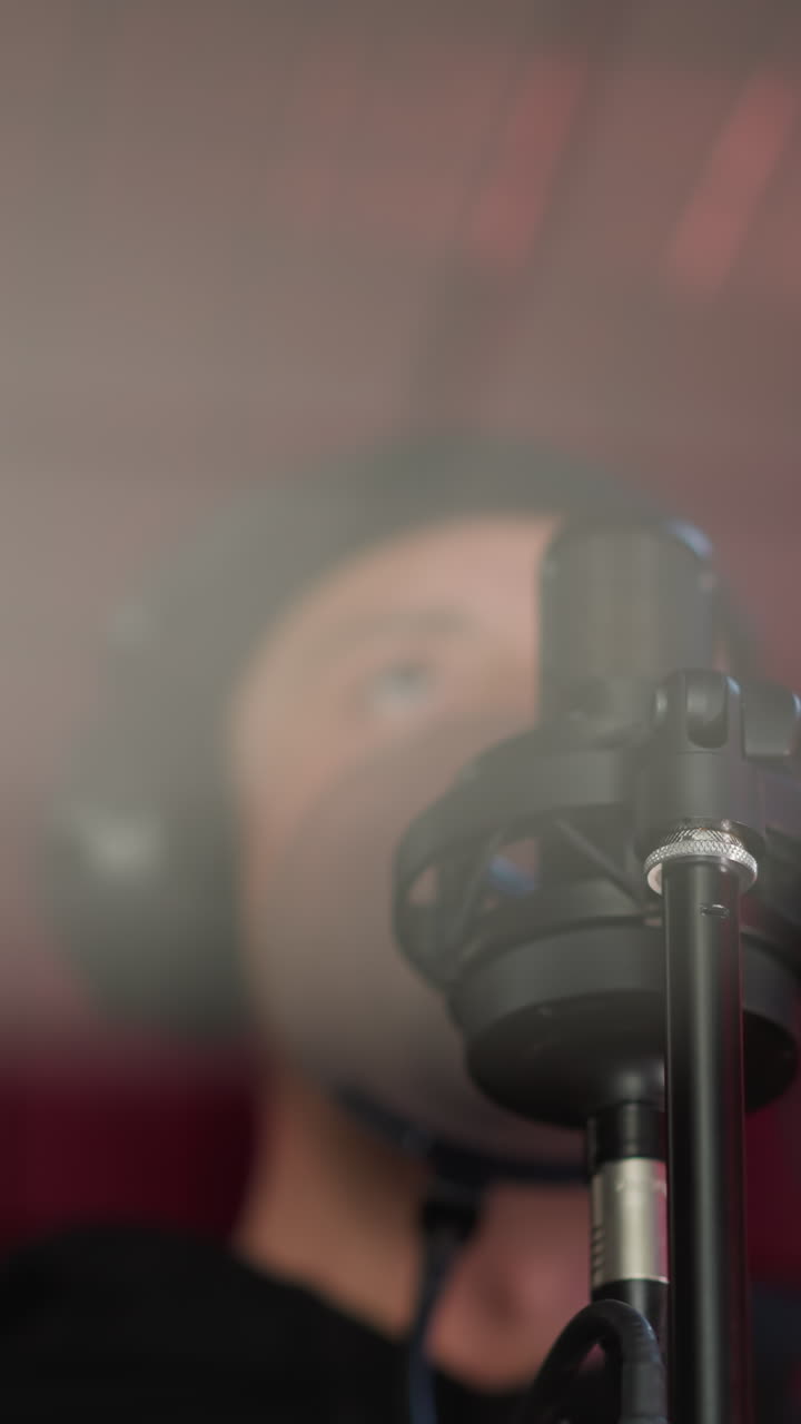 una toma borrosa de un hombre vestido de negro, con auriculares y de pie frente a un micrófono en una habitación a prueba de sonido roja. se centra en el micró fono, con el cantante en el fondo