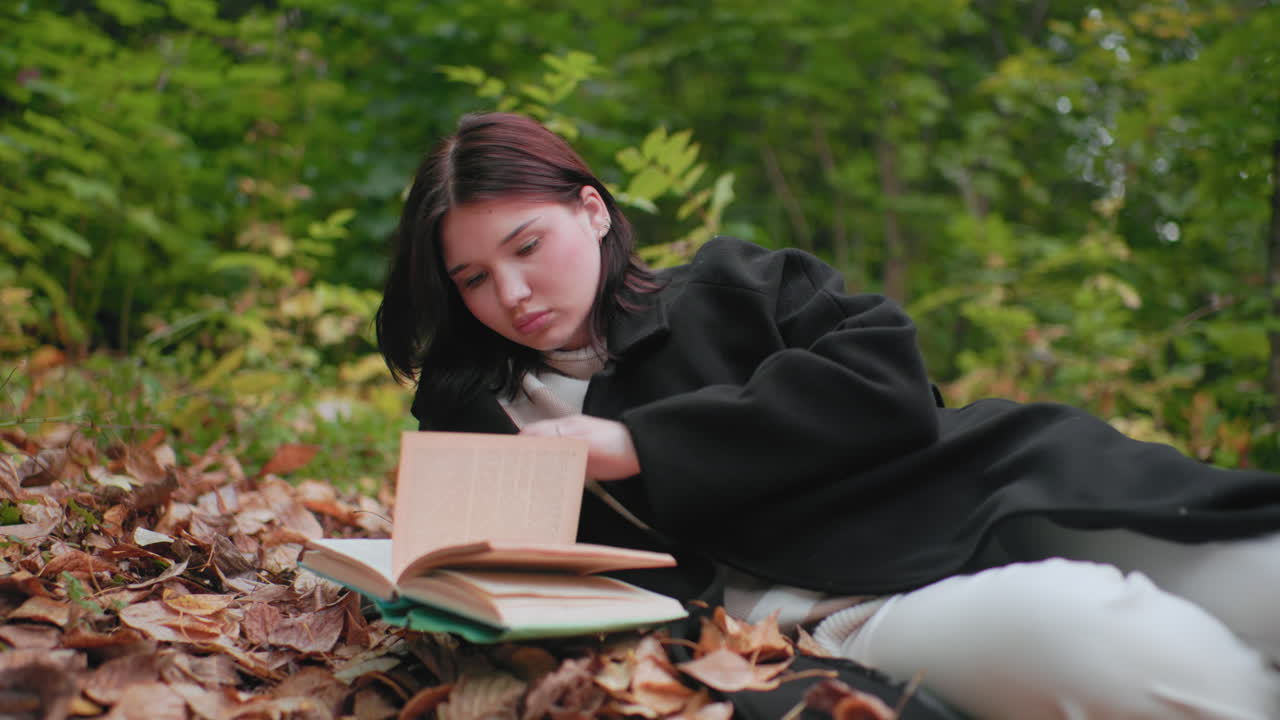 숲 속 낙엽 카펫에 누워 책장을 넘기는 가을을 사랑하는 사람, 몰두한 표정, 아늑한 코트와 흰 바지, 나뭇잎으로 둘러싸인 고요하고 한적한 분위기, 평화로운 독서 시간