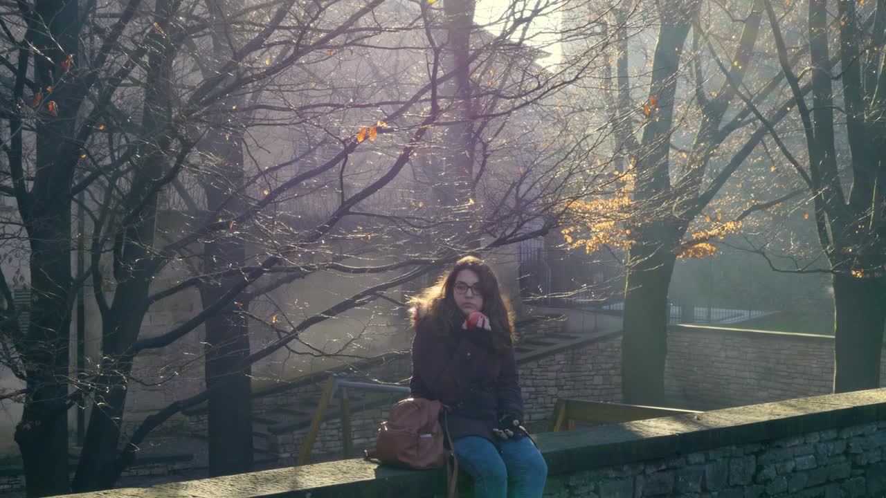 Young woman sitting on an old wall eats an apple while looking at the beautiful park
