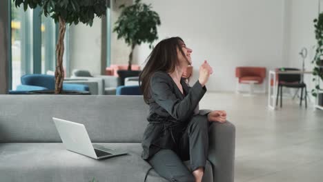 young-tense-woman-is-sitting-on-the-couch-in-the-business-center's-lobby-with-a-laptop,-rejoicing-at-her-victory,-waving-her-hands