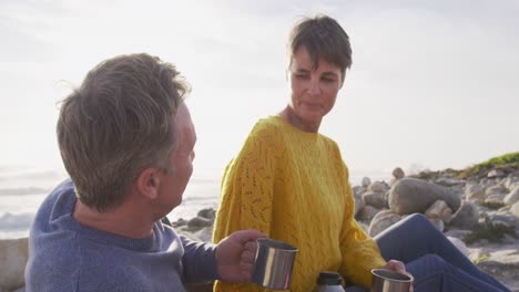 Caucasian-couple-enjoying-free-time-by-sea-on-sunny-day-drinking-coffee