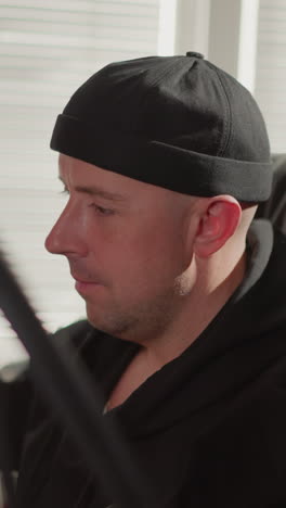 close-up of two men in a studio, with one seated in front of a computer, likely a producer, wearing black clothes and a cap, while the other leans over, gently shaking his head