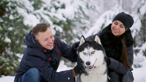 Ein-Mann-Und-Eine-Frau-Sitzen-Und-Umarmen-Einen-Hund,-Einen-Siberian-Husky,-Im-Winterwald,-Lächeln-Und-Schauen-Einander-Und-In-Die-Kamera.-Zeitlupe