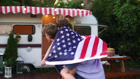 Carefree,-cheerful-couple-having-fun-near-trailer-in-the-park,-man-piggybacking-girl-with-American-flag-on-her-back---spinning-her-around.-Slow-motion