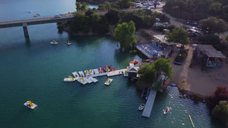 Aerial-view-of-the-jetty-with-boats,-kayaks,-green-trees-around-and-the-bridge-in-the-background