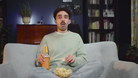 young man watching unexpected movie scene on tv while eating chips and drink soda sitting with crossed legs on couch at home 1