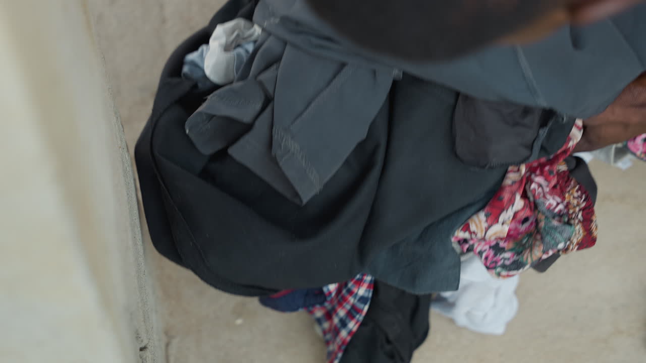 Pila di biancheria scura sul pavimento, un bambino che piega i vestiti vicino a scale di cemento all'interno di una casa, mani che smistano asciugamani e magliette, tonalità tenui e tessuto texturizzato, un'angolazione dall'alto spontanea che trasmette una tranquilla atmosfera domestica.