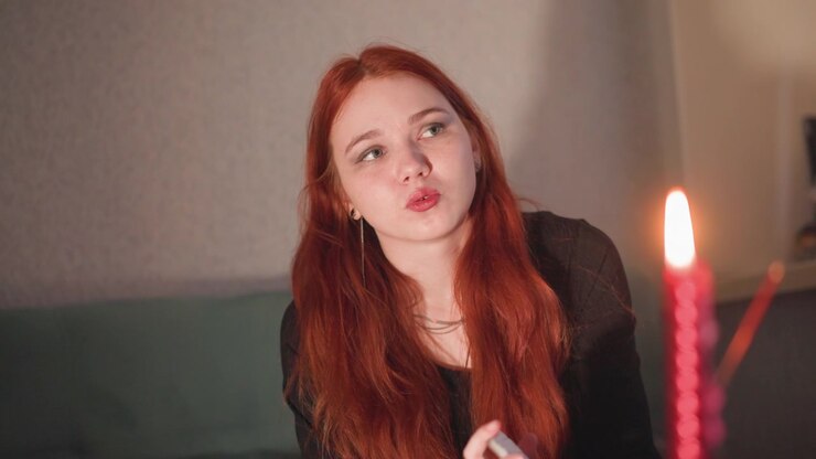 Closeup redhead seer by candle, young oracle leans forward breathing toward flame, lips pursed and eyes searching, warm candlelight highlights freckled skin, soft indoor background, introspective