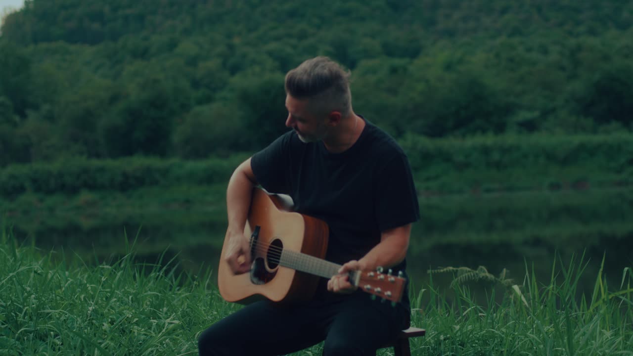 Mann spielt Akustikgitarre an einem Fluss im Wald