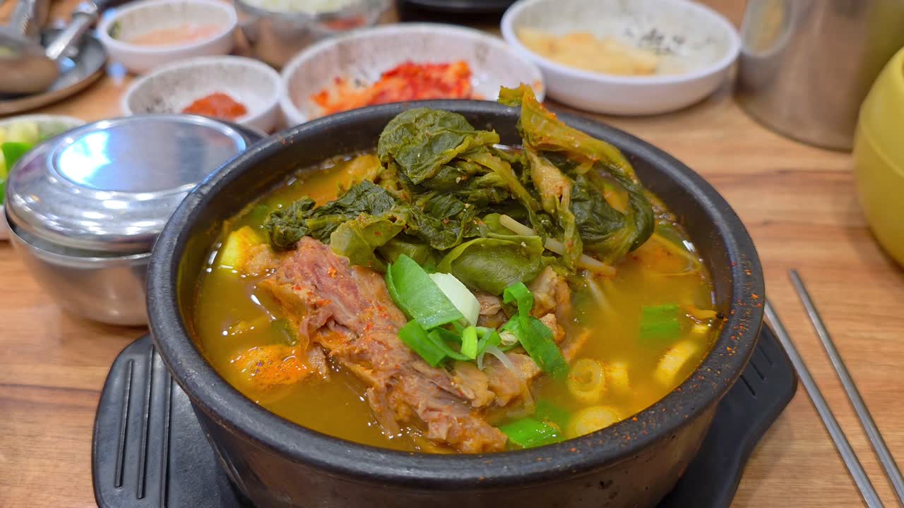A steaming bowl of Korean pork bone soup (gamjatang) topped with greens, served with rice, kimchi, and assorted side dishes on a wooden table in a traditional restaurant setting - slow dolly in
