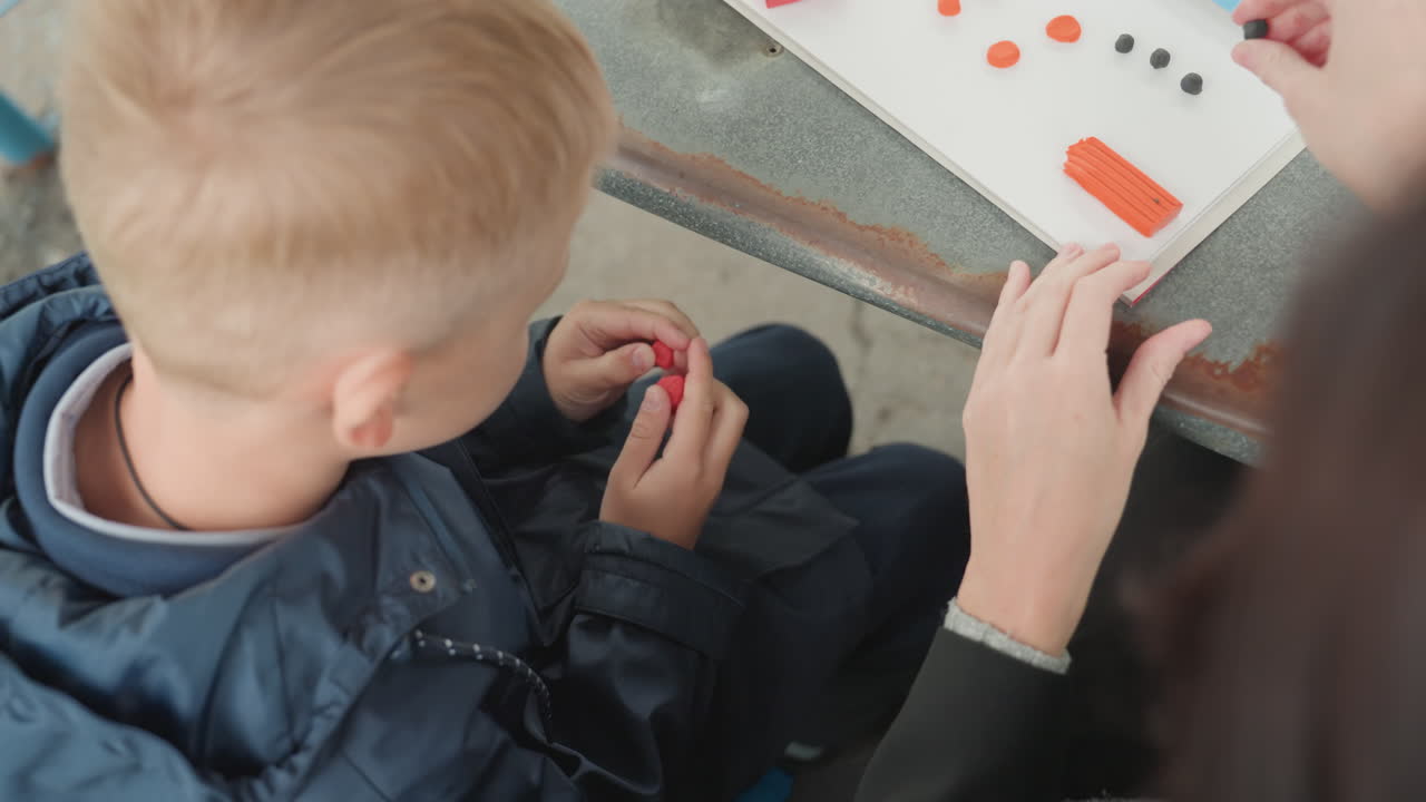 Ragazzo caucasico che prepara un lavoretto scolastico all'aperto. Un adulto di riferimento guida il lavoro con piccole perline rosse, mani concentrate sul tavolo, bambino con la giacca che si concentra sulla manipolazione tattile e sulla pratica delle abilità.