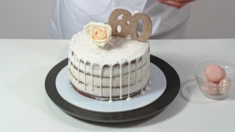 beautiful birthday cake, decorated with macarons and a rose, being proudly presented by a female baker.