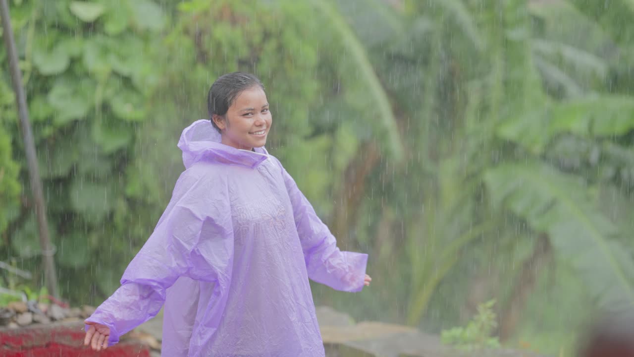 Young Indian girl in raincoat spreading her arms and smiling while standing in the rain, countryside scene, 4k video