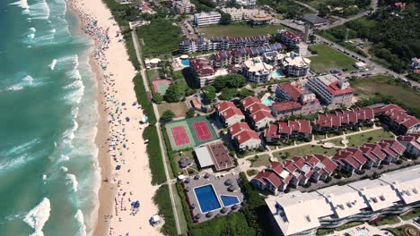 Panoramic aerial view of Brava beach in Florianopolis with many people on the sand, surrounded by mountains and vegetation with urban area, buildings and houses by the sea - orbital scene