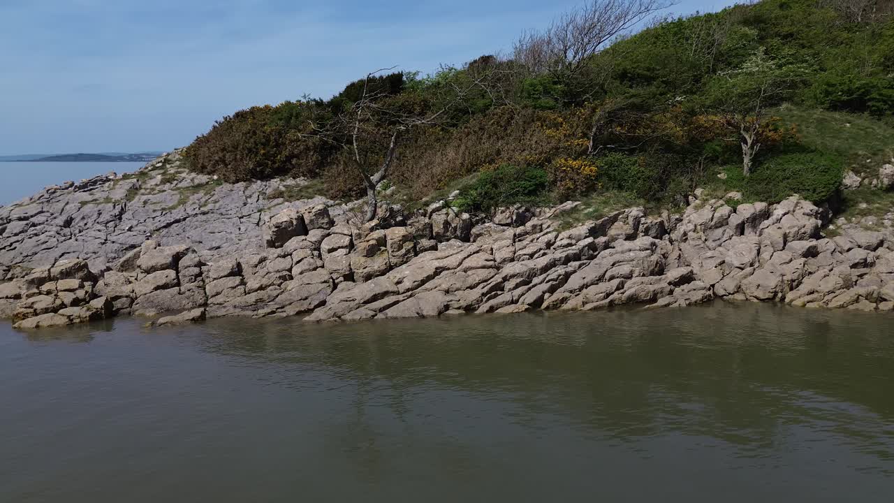 Aerial view circling steep rocky shoreline with weathered tree and reflections in calming tide