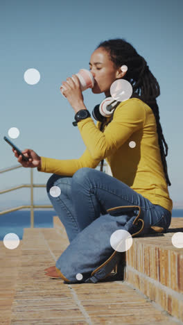 white spots against african american woman drinking coffee and using smartphone on the promenade