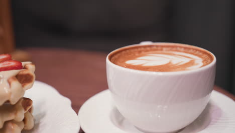 close-up view of latte with leaf-shaped milk foam art in white cup on saucer, placed beside stack of waffles topped with strawberries and cream on wooden table in cozy setting