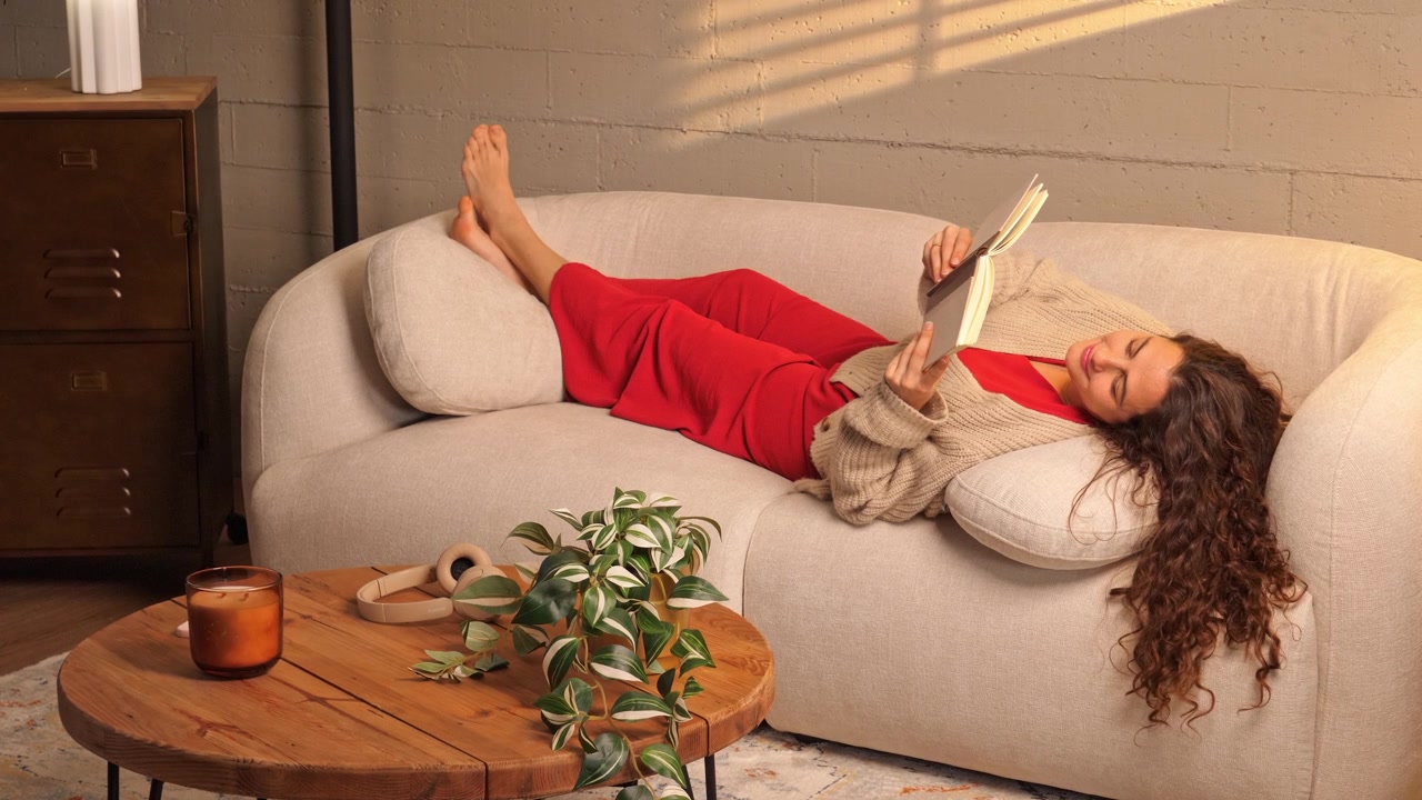 Ampia inquadratura di una donna rilassata sdraiata su un divano che legge un libro in un soggiorno elegante e accogliente con tavolino da caffè e pianta. Concetto per stile di vita confortevole, interior design e comfort domestico