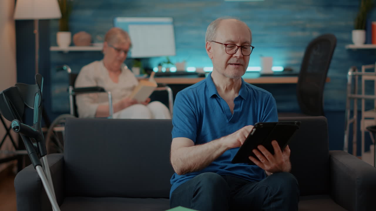 Senior Man Using Tablet with Elderly Woman Reading in Background