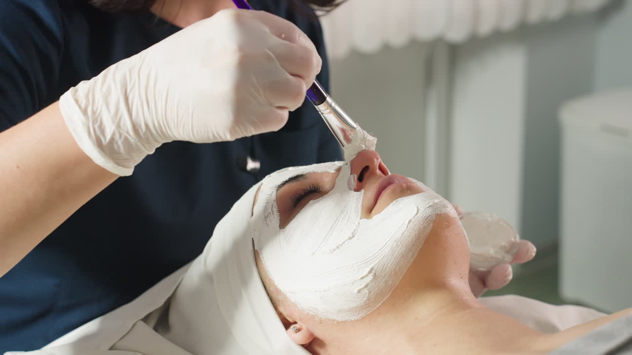 Treatment Provider Applies Face Mask Carefully With Soft Silicone Brush On Customer Skin During Relaxing Spa Session Under Bright Studio Light Highlighting Professional Technique