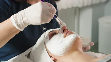 treatment provider applies face mask carefully with soft silicone brush on customer skin during relaxing spa session under bright studio light highlighting professional technique