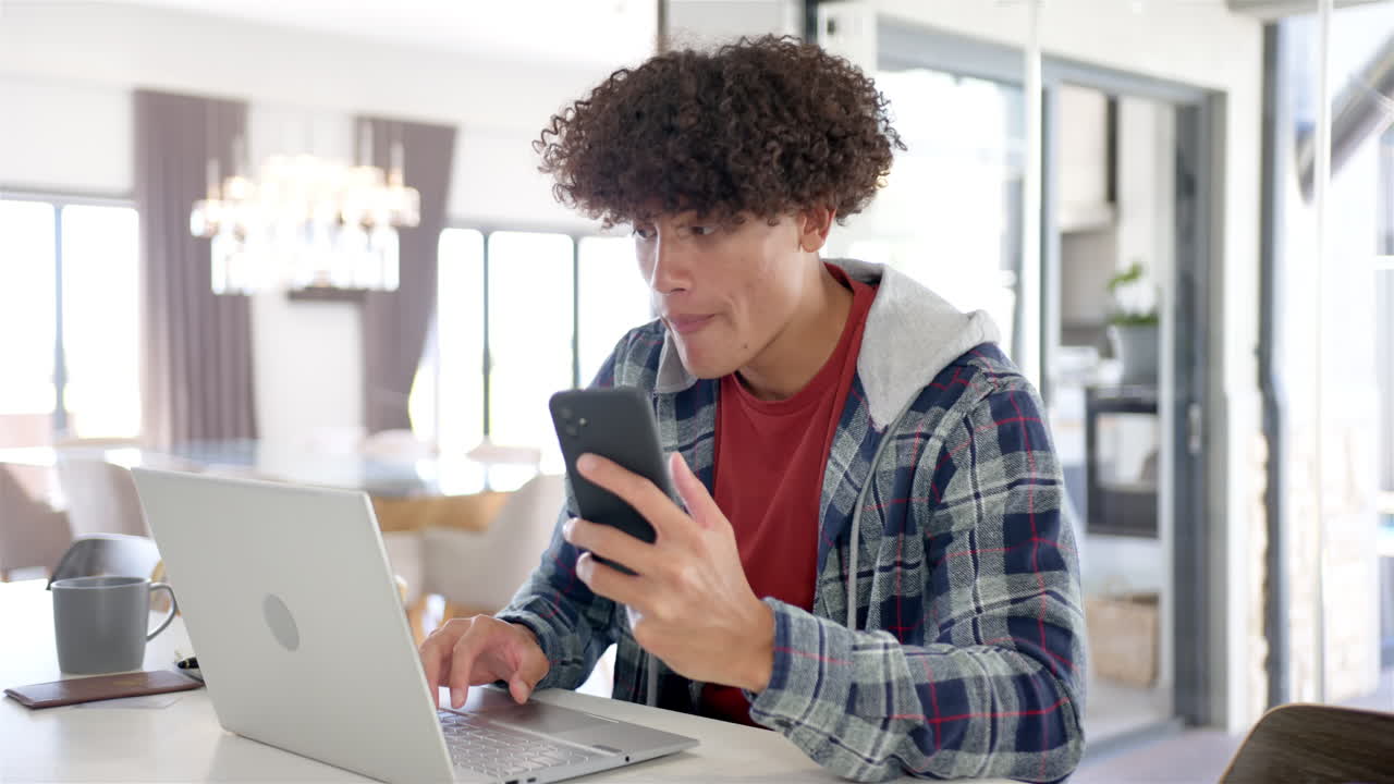 A young biracial man is using his smartphone and laptop at home