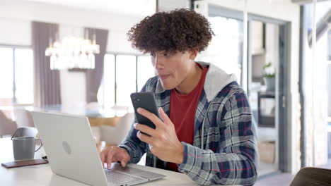 un joven biracial está usando su teléfono inteligente y portátil en casa