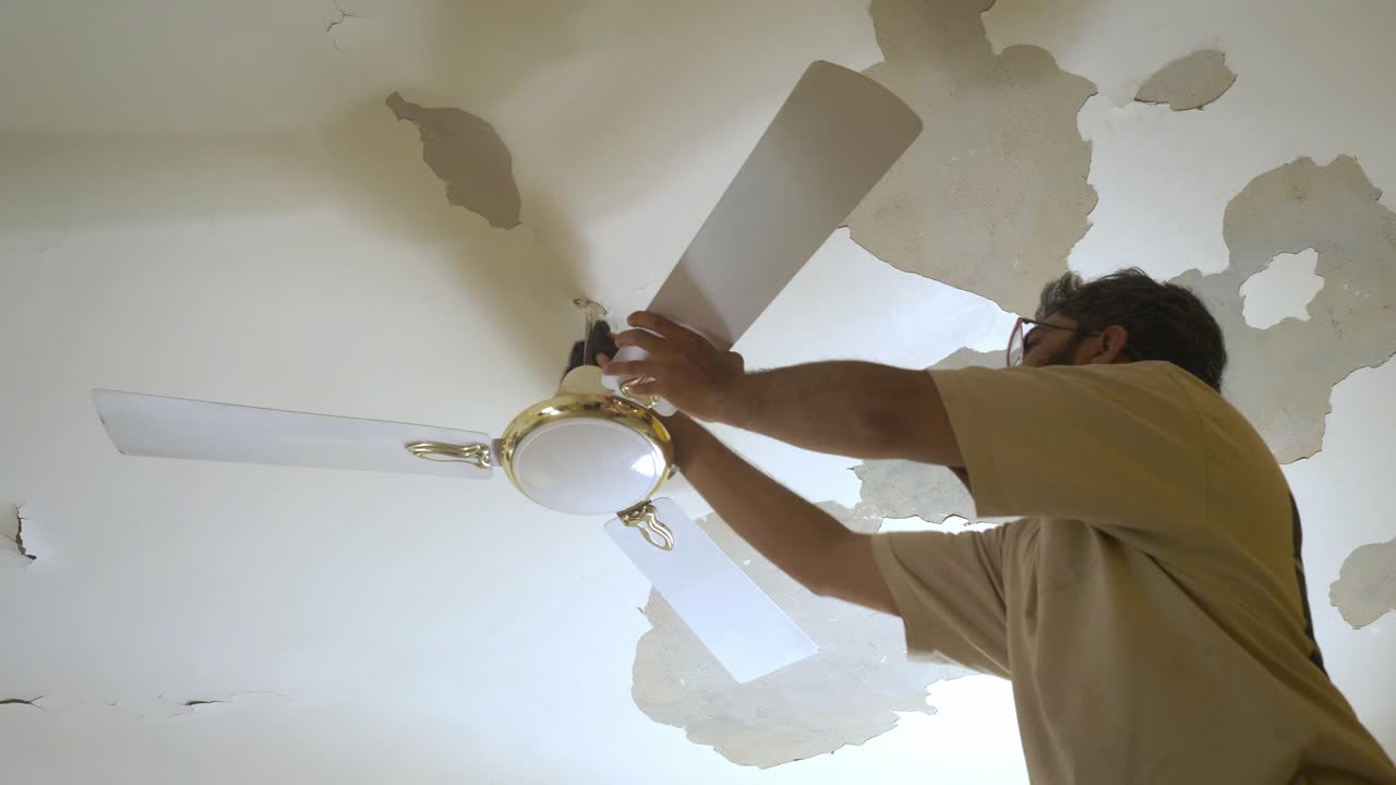 South Asian man cleaning ceiling fan of bedroom