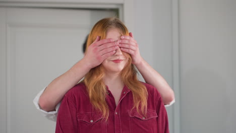 woman in red shirt stands calmly with eyes closed as man in sunglasses and black cap gently covers her eyes from behind, preparing to lead her forward with a playful gesture in bright indoor space