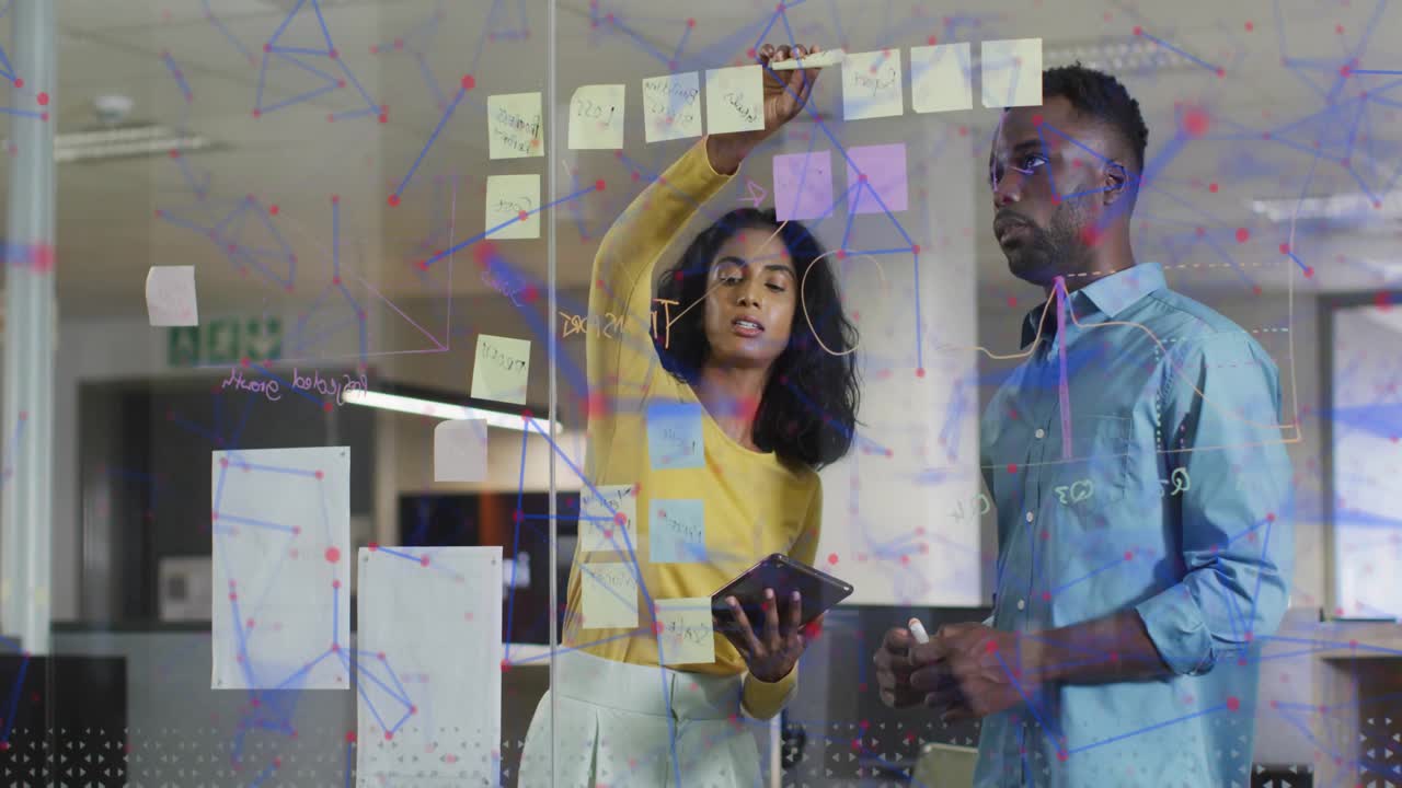 Woman tapping tablet, placing pink note on glass board as male colleague pointing for business plan