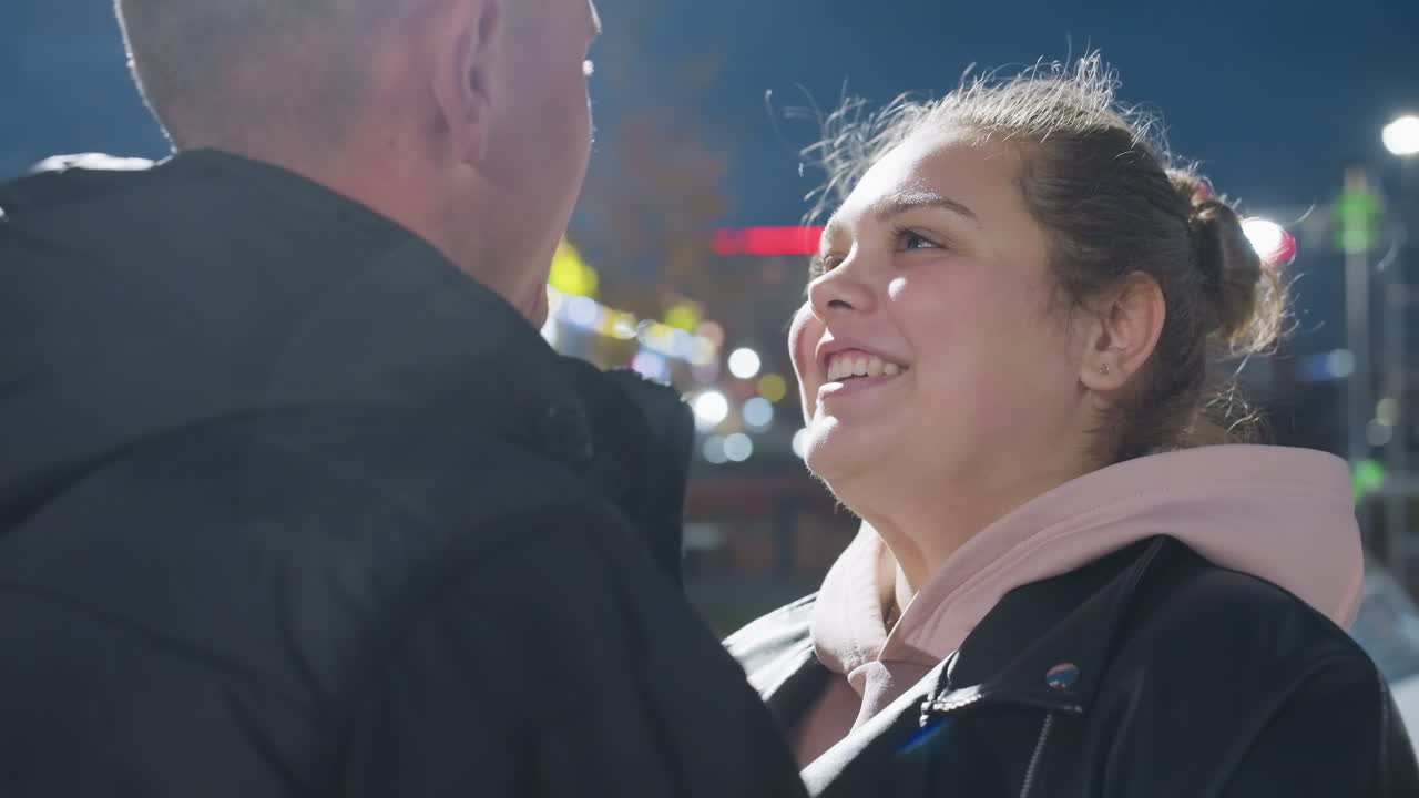 Close up of lady and man laughing closely while holding each other warmly with blurred city lights glowing in background under night sky capturing joyful connection