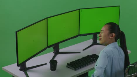 vista lateral de una mujer asiática con dolor de espalda mientras trabaja con múltiples monitores de computadora simulados en un estudio de pantalla verde