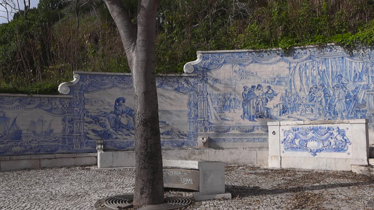 Lenta panoramica su una fontana storica di piastrelle azulejo e un piccolo parco costiero a Paco de Arcos, con opere d'arte in ceramica bianca e blu e pavimentazione in calcada portuguesa in una giornata di sole