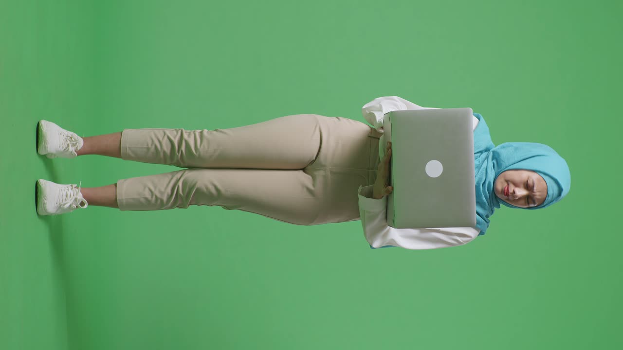 cuerpo lleno de mujer musulmana asiática escribiendo en una computadora portátil y teniendo dolor de cabeza mientras estaba de pie en el estudio de fondo de pantalla verde