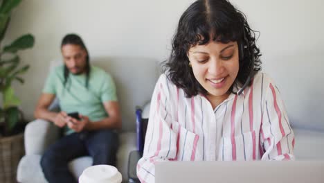 Happy-biracial-woman-in-wheelchair-using-laptop-and-male-partner-using-smartphone-in-living-room