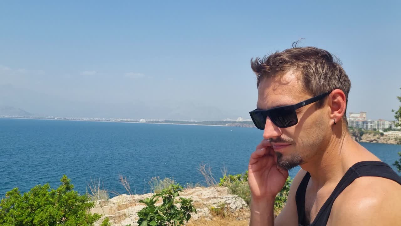 Close-up of a man in sunglasses scratching his cheek while sitting on a cliff above the Mediterranean Sea near Antalya, Turkey